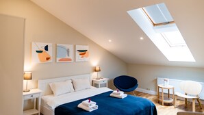 A modern bedroom with a sloped ceiling, a skylight, a bed with white linens and a blue blanket, two bedside tables with lamps, and abstract wall art.