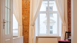 A room with a brick wall, a radiator, a window with white curtains, a wooden floor, a white cabinet, and a chair.