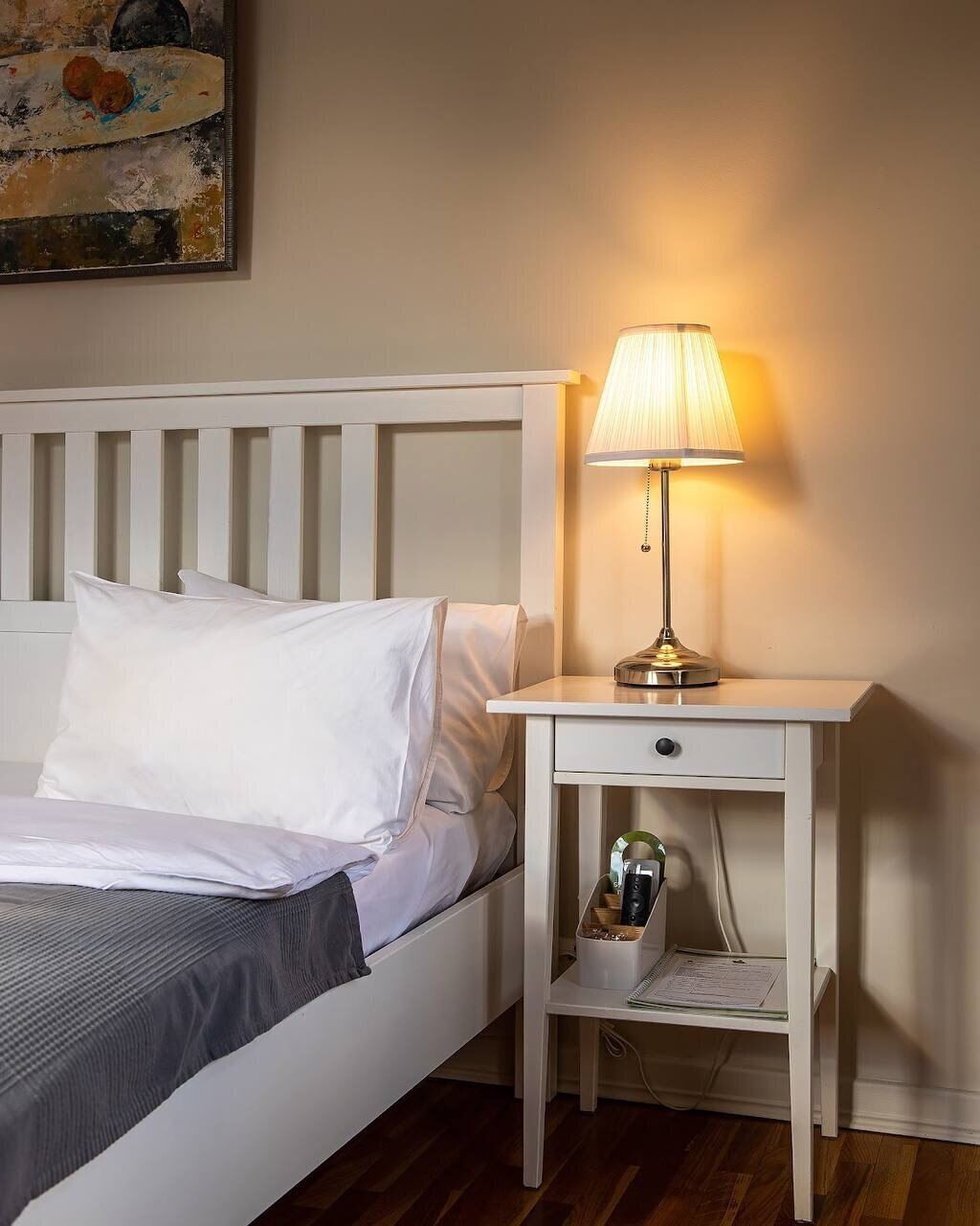 A neatly made bed with white linens, a bedside table with a lamp, and a small drawer unit.