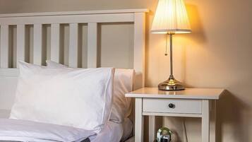 A neatly made bed with white linens, a bedside table with a lamp, and a small drawer unit.
