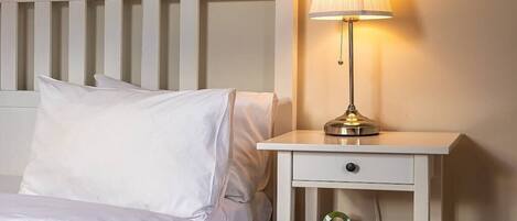 Una cama bien tendida con sábanas blancas, una mesita de noche con lámpara y una cómoda pequeña.