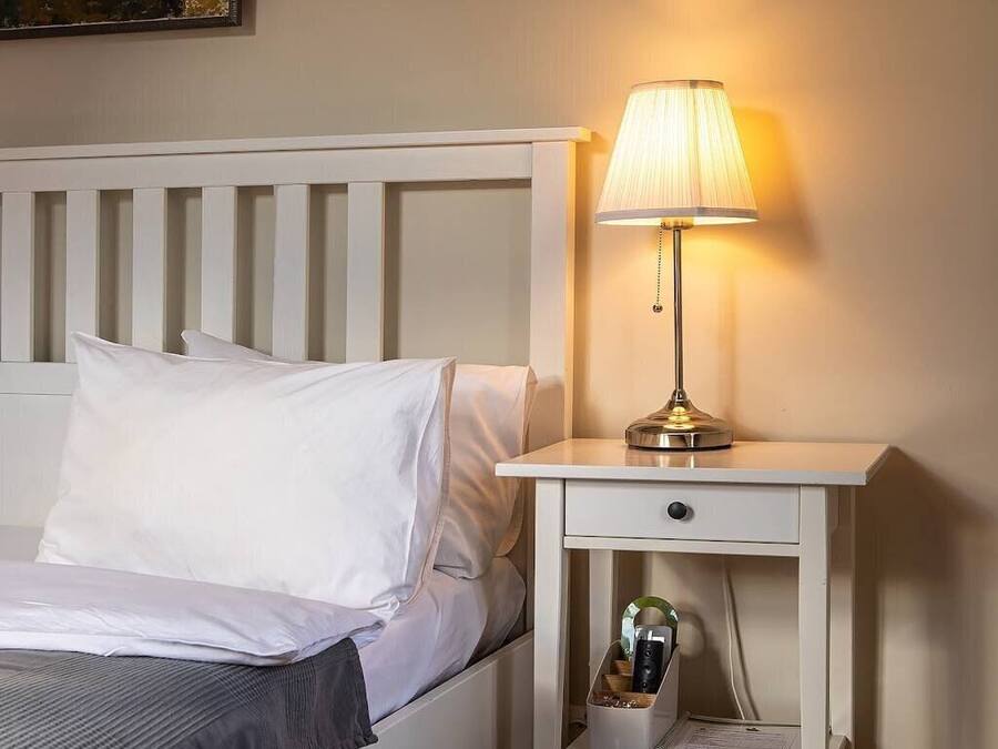 A neatly made bed with white linens, a bedside table with a lamp, and a small drawer unit.