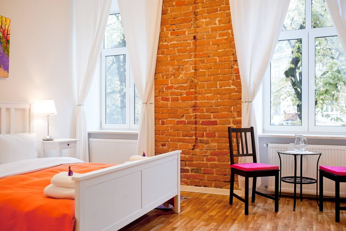 A bedroom with a brick wall, a bed with an orange bedspread, a chair, and a small table with a pitcher.