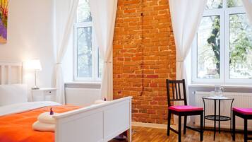 A bedroom with a brick wall, a bed with an orange bedspread, a chair, and a small table with a pitcher.