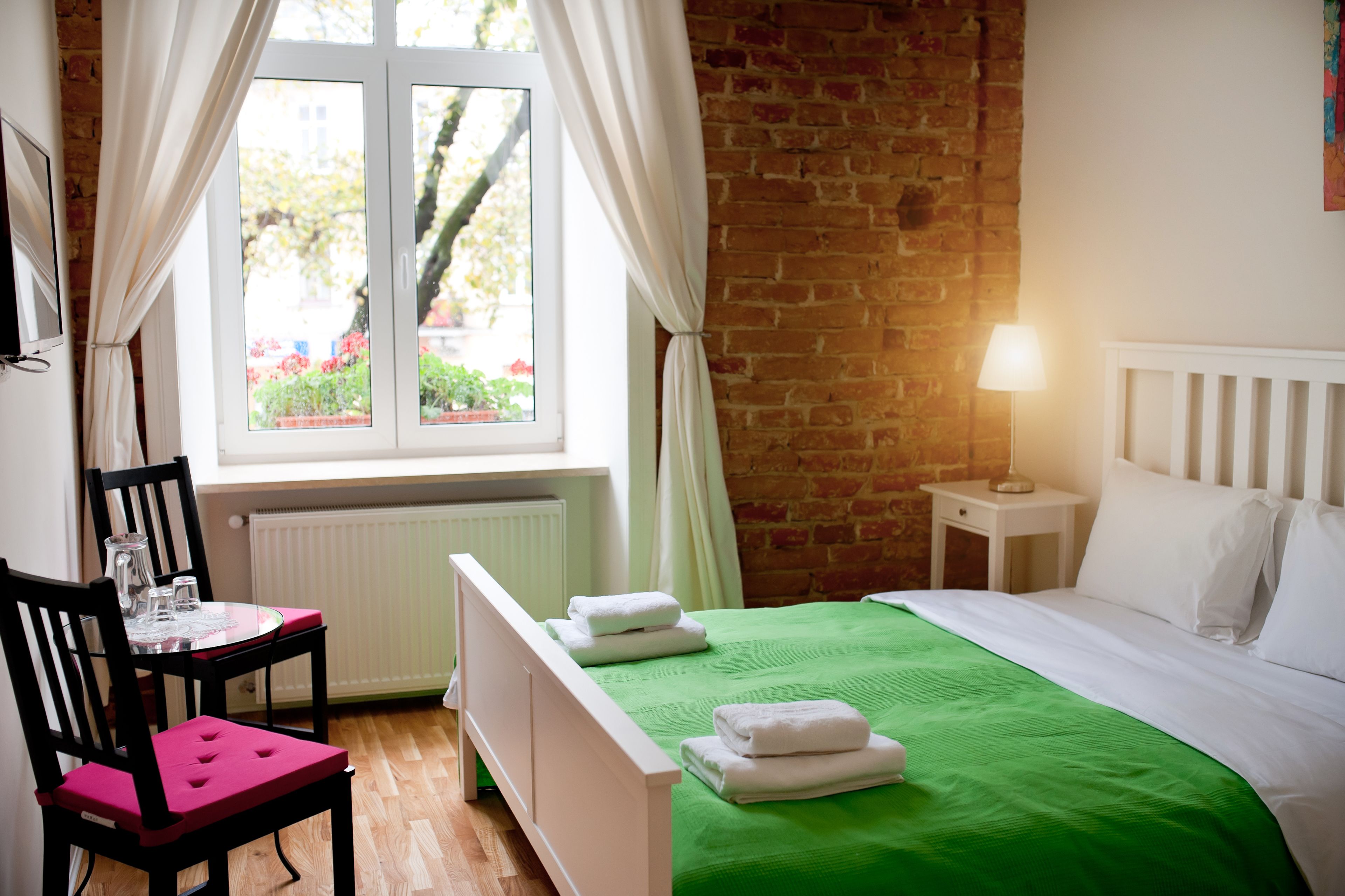 Ein Zimmer mit Backsteinwand, einem Bett mit grüner Bettdecke, einem weißen Heizkörper, einem kleinen Tisch mit Stuhl und einem Fenster mit Vorhängen.