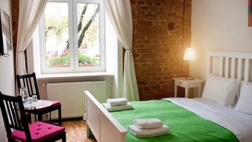 Ein Zimmer mit Backsteinwand, einem Bett mit grüner Bettdecke, einem weißen Heizkörper, einem kleinen Tisch mit Stuhl und einem Fenster mit Vorhängen.
