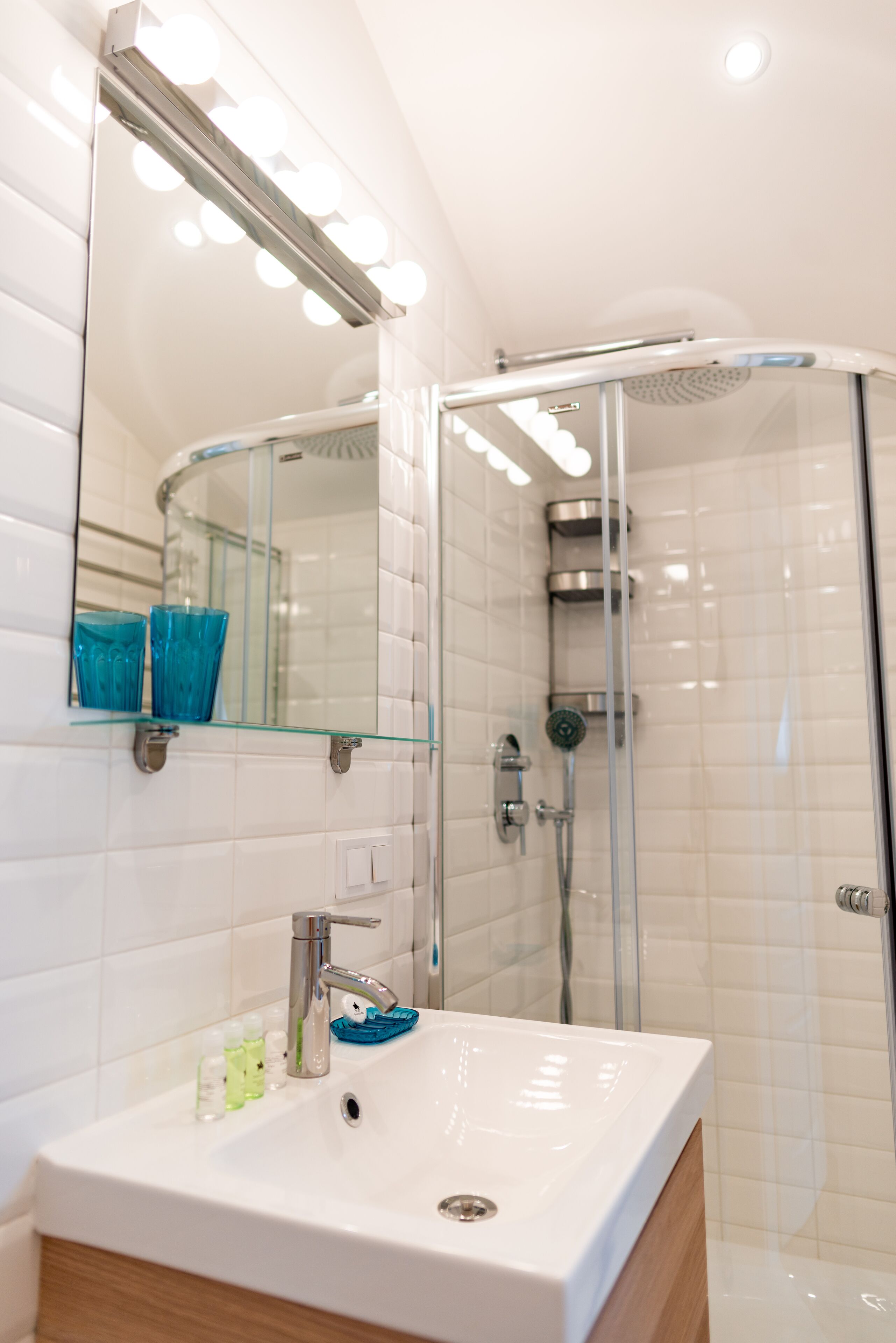 A modern bathroom with a glass shower enclosure, a white sink with a mirror above it, and a glass shelf with toiletries.