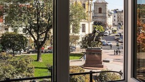 A view from a window overlooking a city street with historic buildings, a statue, and a clear blue sky.