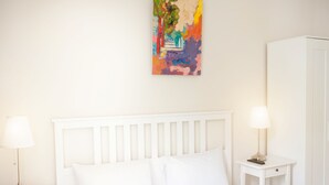 A bedroom with a white bed, two bedside tables with lamps, a white wardrobe, and a colorful abstract painting on the wall.