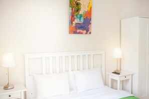 A bedroom with a white bed, two bedside tables with lamps, a white wardrobe, and a colorful abstract painting on the wall.