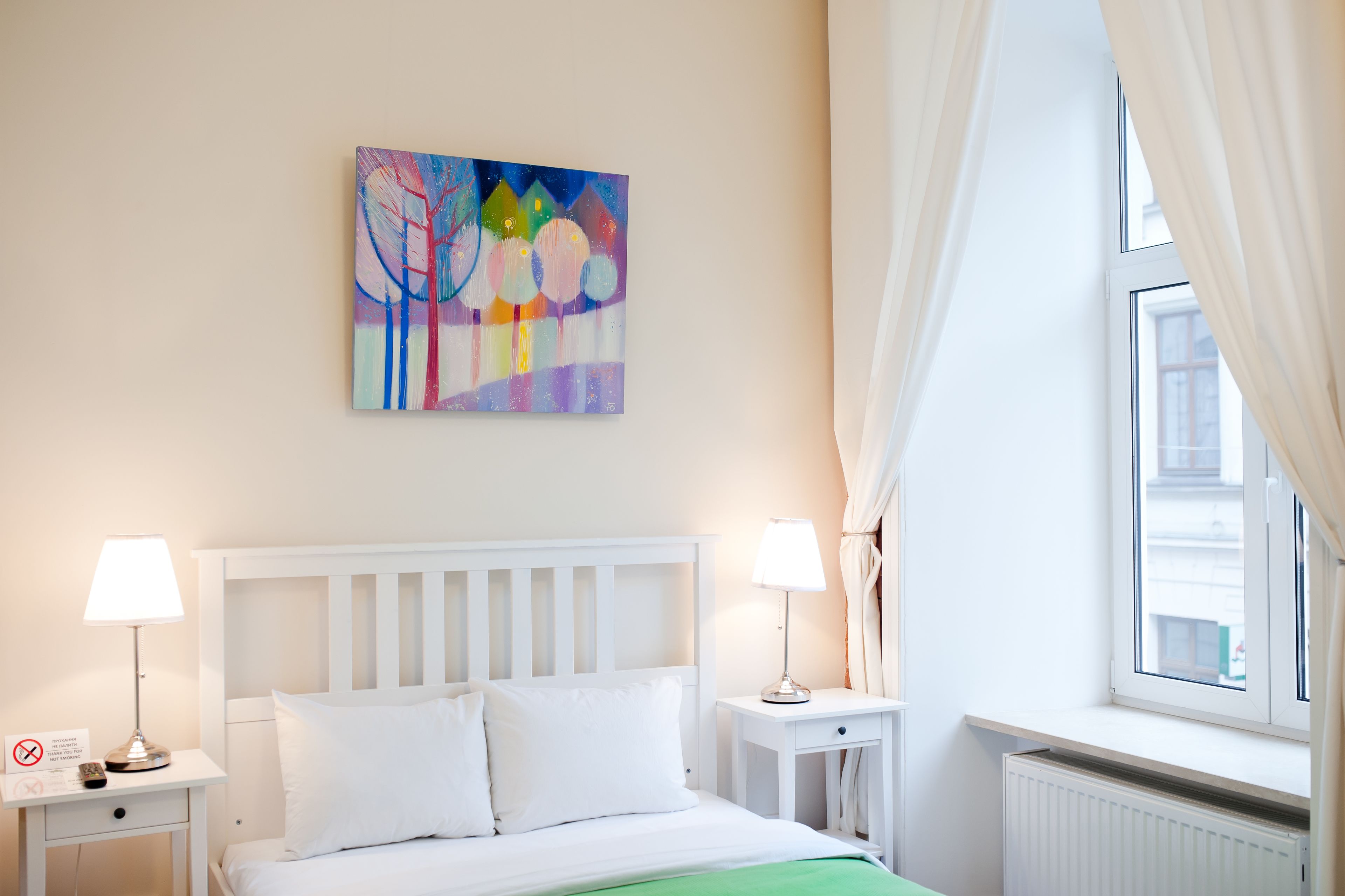 Ein Hotelzimmer mit einem Bett, zwei Nachttischen, einem Heizkörper, einem Fenster mit Vorhängen und einem farbenfrohen Bild an der Wand.