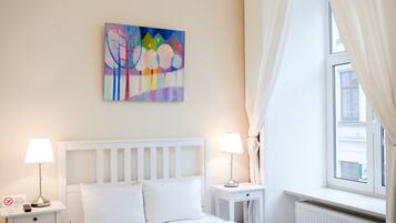 Ein Hotelzimmer mit einem Bett, zwei Nachttischen, einem Heizkörper, einem Fenster mit Vorhängen und einem farbenfrohen Bild an der Wand.