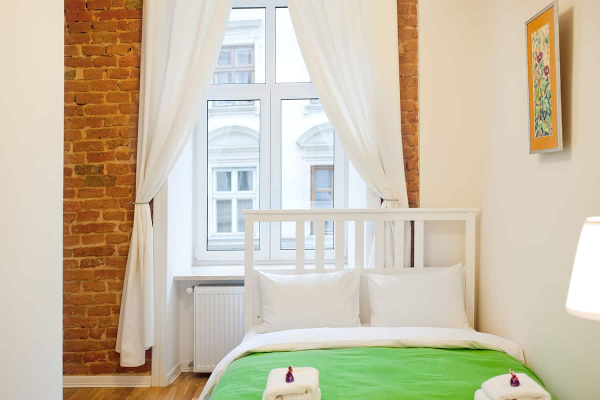 A hotel room with a brick wall, a white bed with green bedding, white curtains, and a window with a view of buildings.