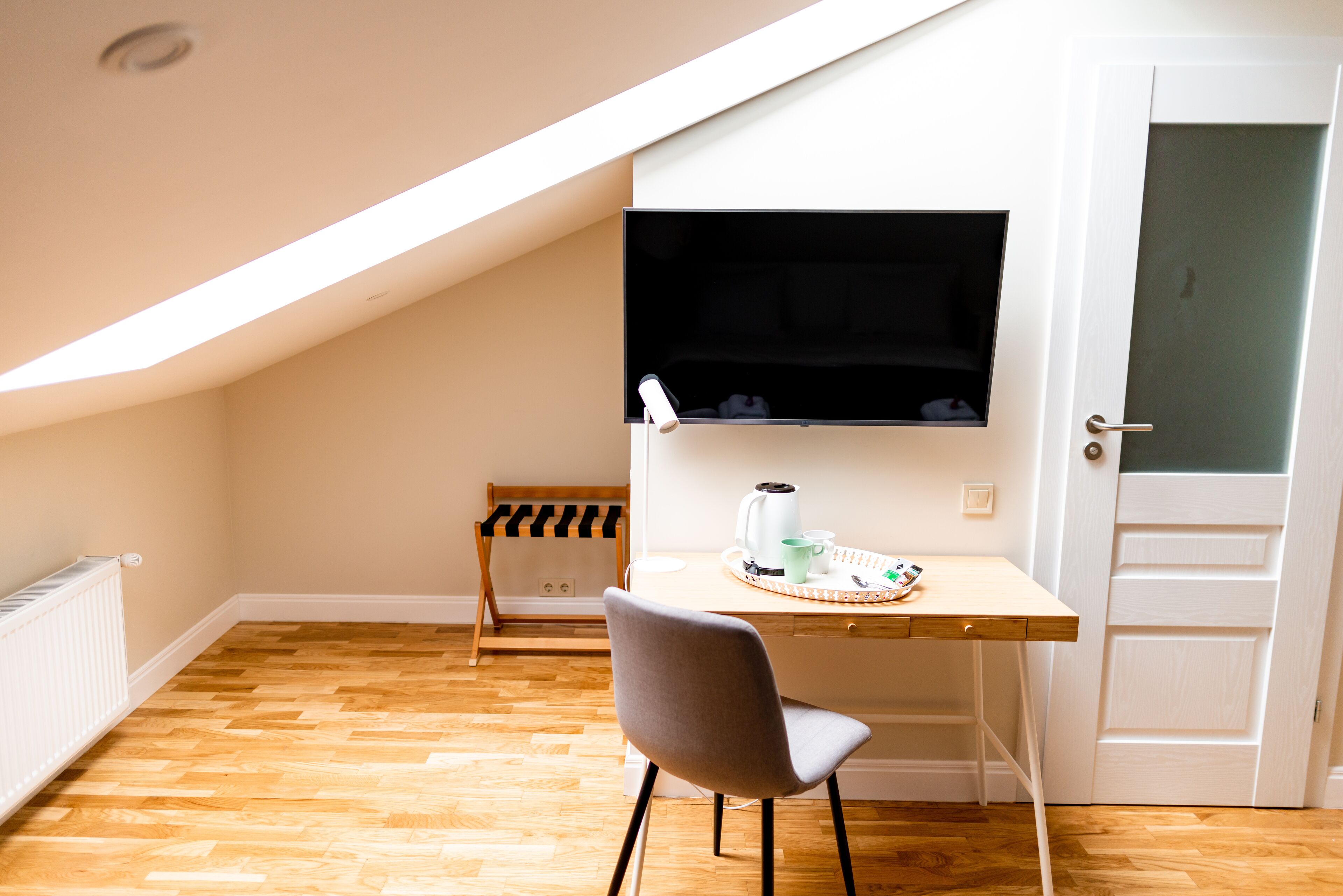 A small, bright room with a sloped ceiling, a wooden floor, a desk with a chair, a television, and a door.