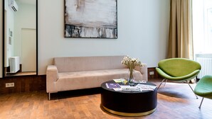 A modern living room with a beige sofa, a round black coffee table with magazines, a green chair, and a large abstract painting on the wall.