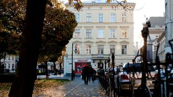 A cobblestone pathway lined with trees and parked cars, leading to a building with a sign that reads "OBNOVOV".
