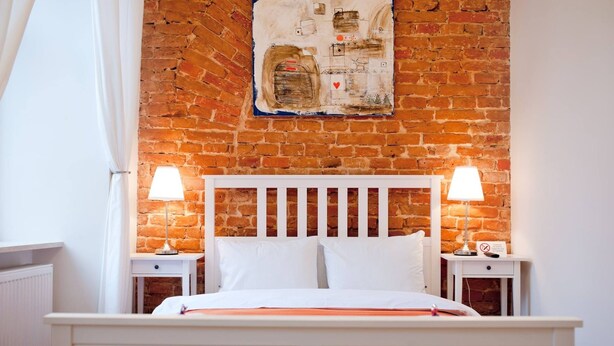 A bedroom with a brick wall, white headboard, two bedside tables with lamps, and a white bed.
