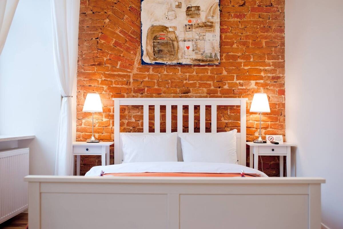 A bedroom with a brick wall, a white headboard, two bedside tables with lamps, and a bed with white bedding.