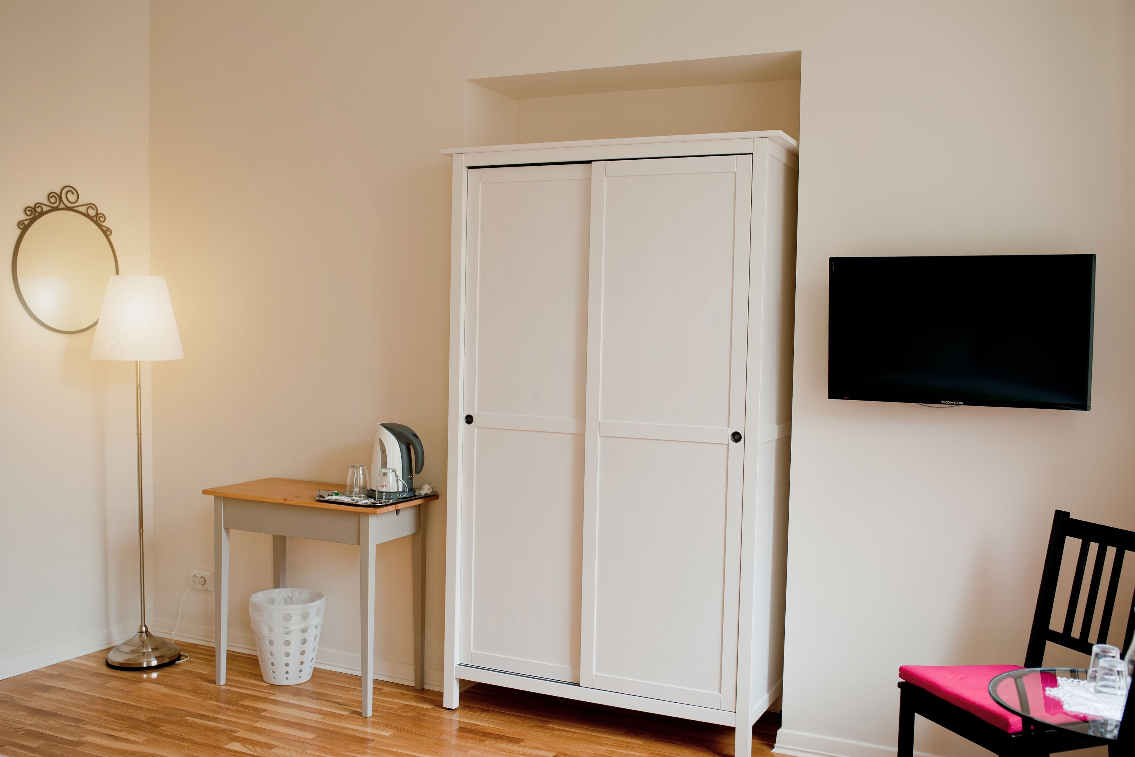 Une pièce avec une armoire blanche, une petite table avec une lampe, une chaise et un téléviseur fixé au mur.