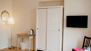 Une pièce avec une armoire blanche, une petite table avec une lampe, une chaise et un téléviseur fixé au mur.