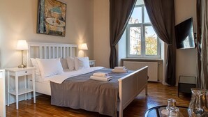 A neatly arranged bedroom with a bed, bedside lamps, a painting, a window with curtains, and a television.