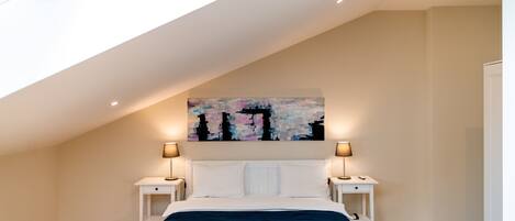 Un dormitorio con techo inclinado, una cama grande con ropa de cama blanca, dos mesitas de noche con lámparas y un cuadro abstracto y colorido en la pared.