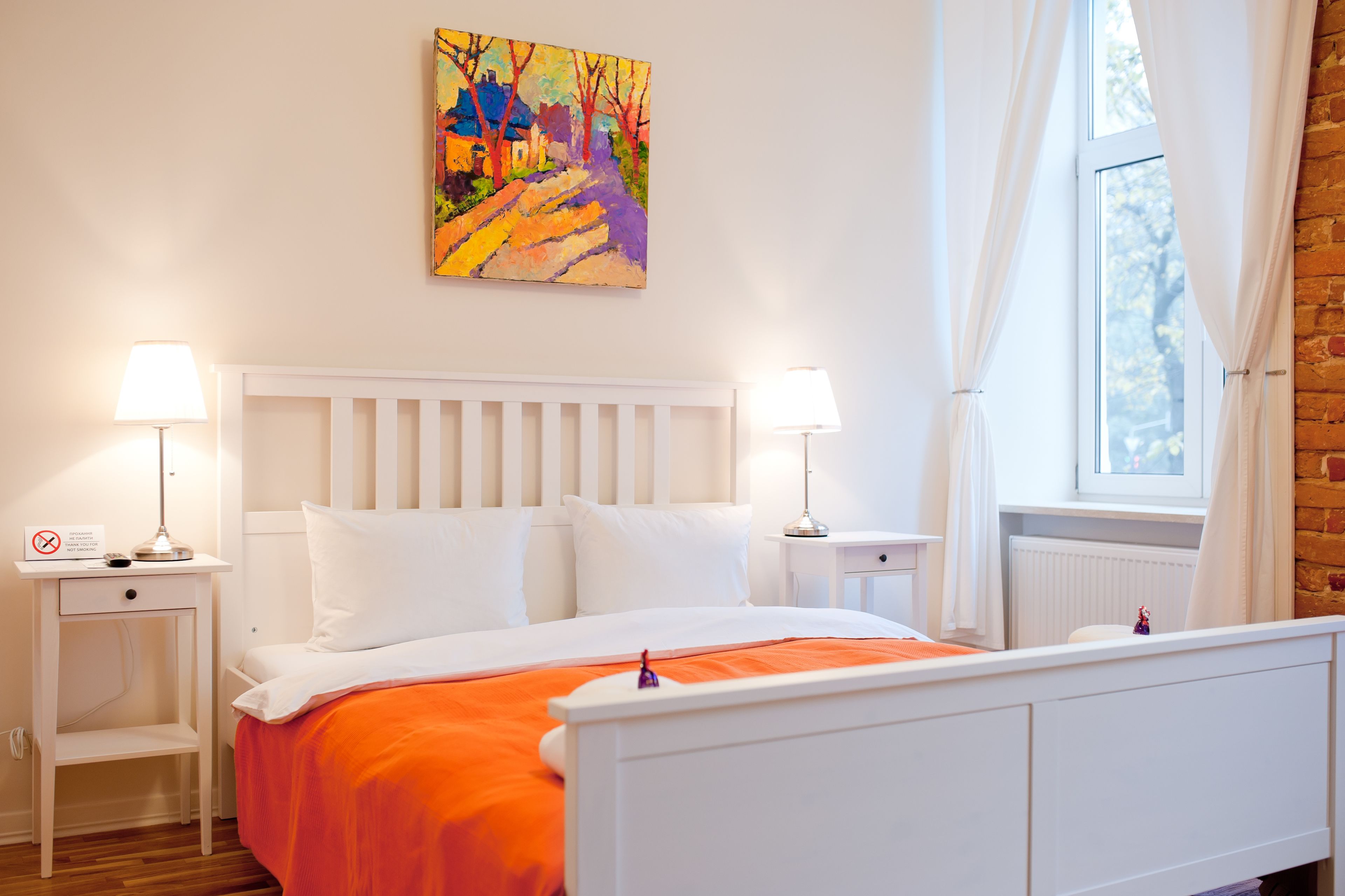 Un dormitorio con una cama blanca, ropa de cama naranja, dos mesitas de noche con lámparas, un cuadro en la pared y una ventana con cortinas.