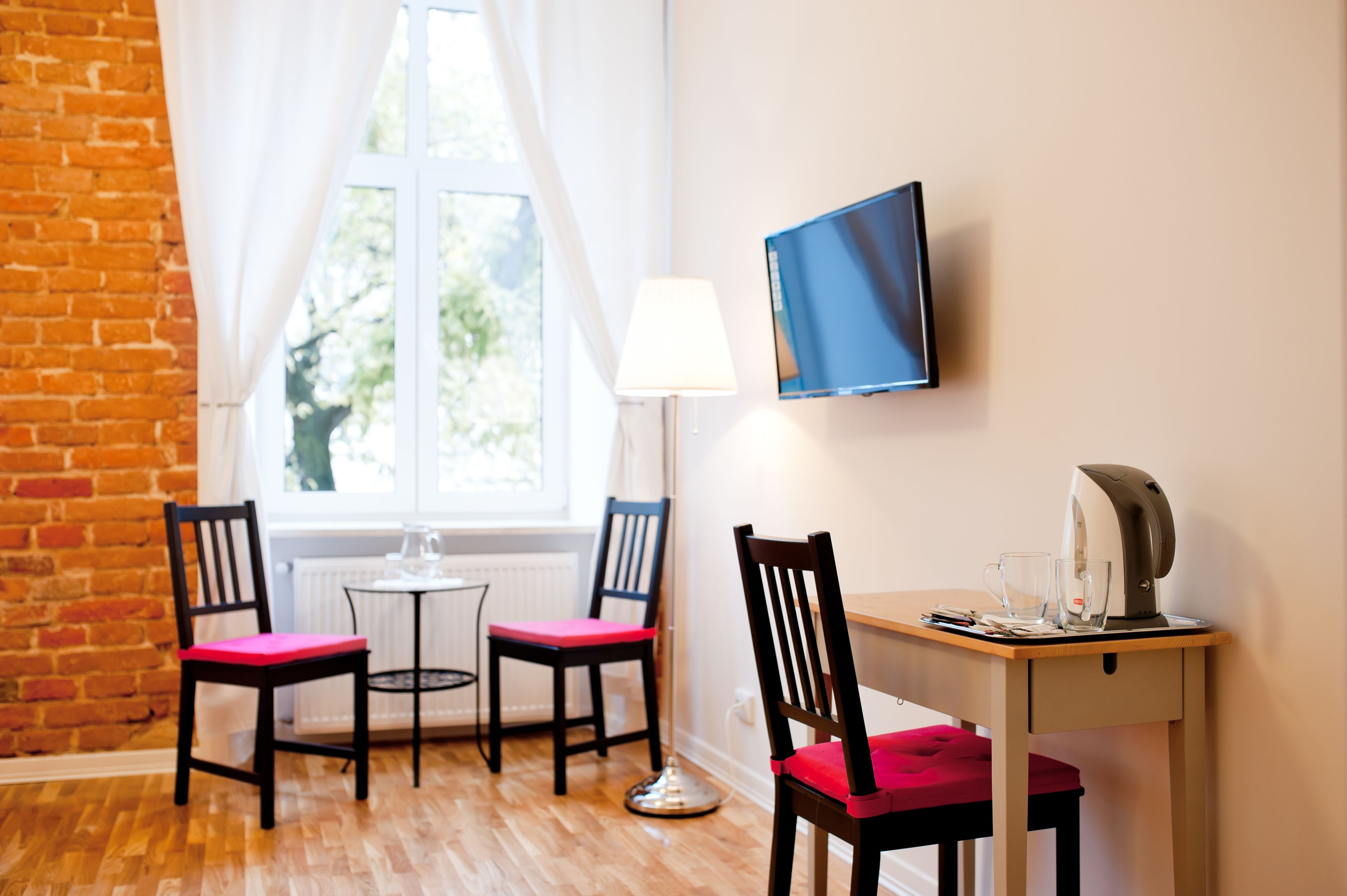 Ein Zimmer mit Backsteinwand, einem Vorhangfenster, einem Schreibtisch mit Computermonitor, zwei Sesseln mit roten Kissen, einer Stehlampe und einer Kaffeemaschine.