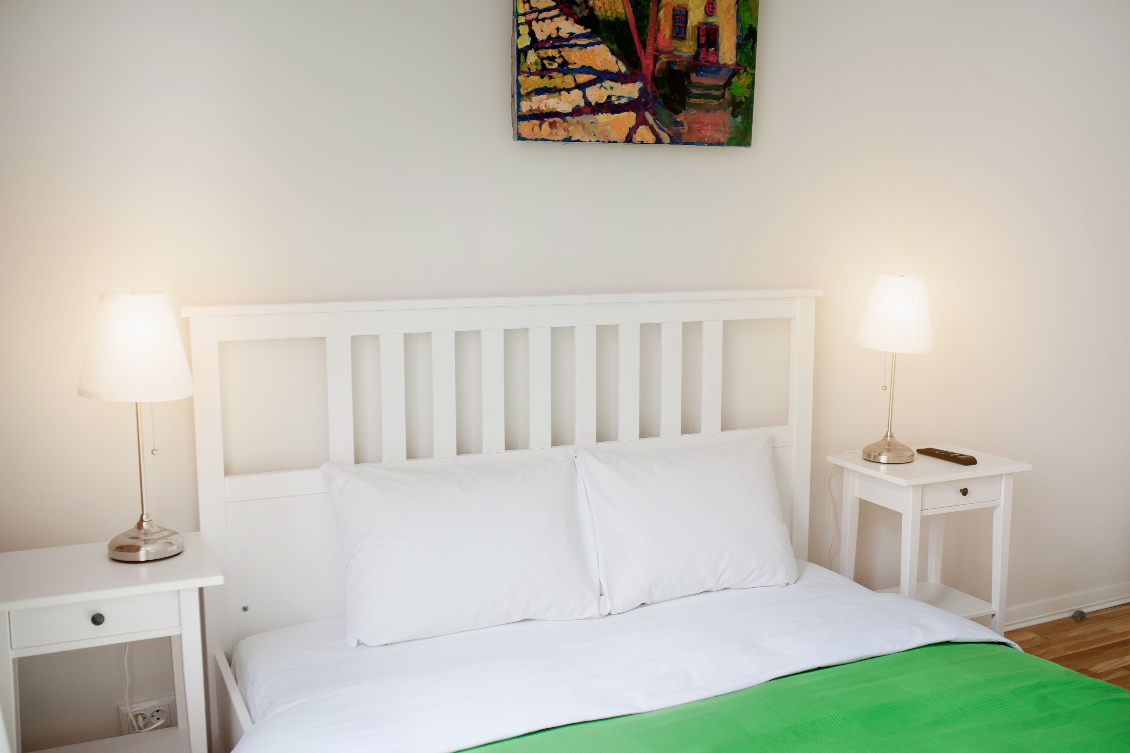 Una cama blanca con una manta verde, dos mesitas de noche con lámparas, y un cuadro en la pared.
