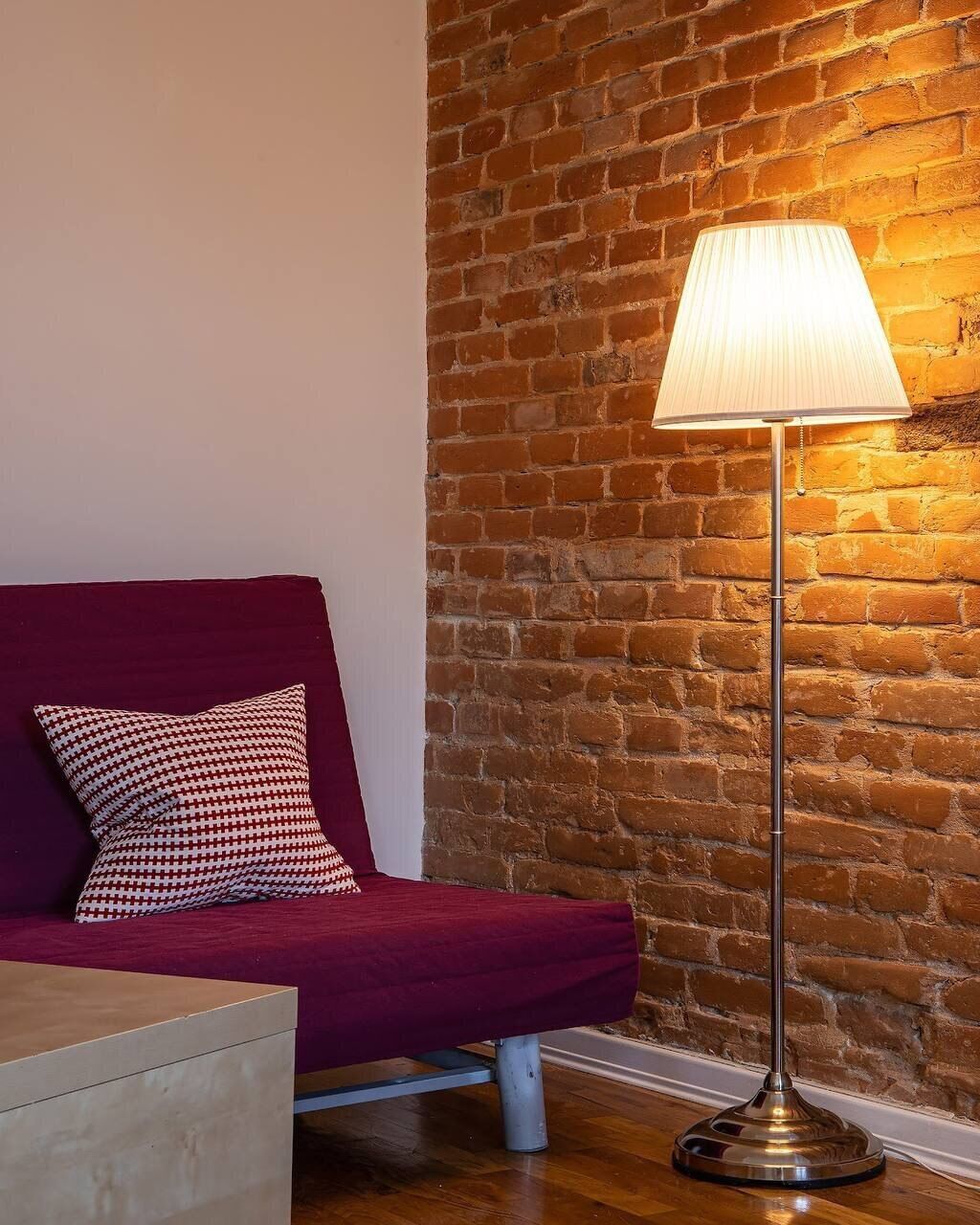 A room with a brick wall, a red sofa, a patterned pillow, and a floor lamp.