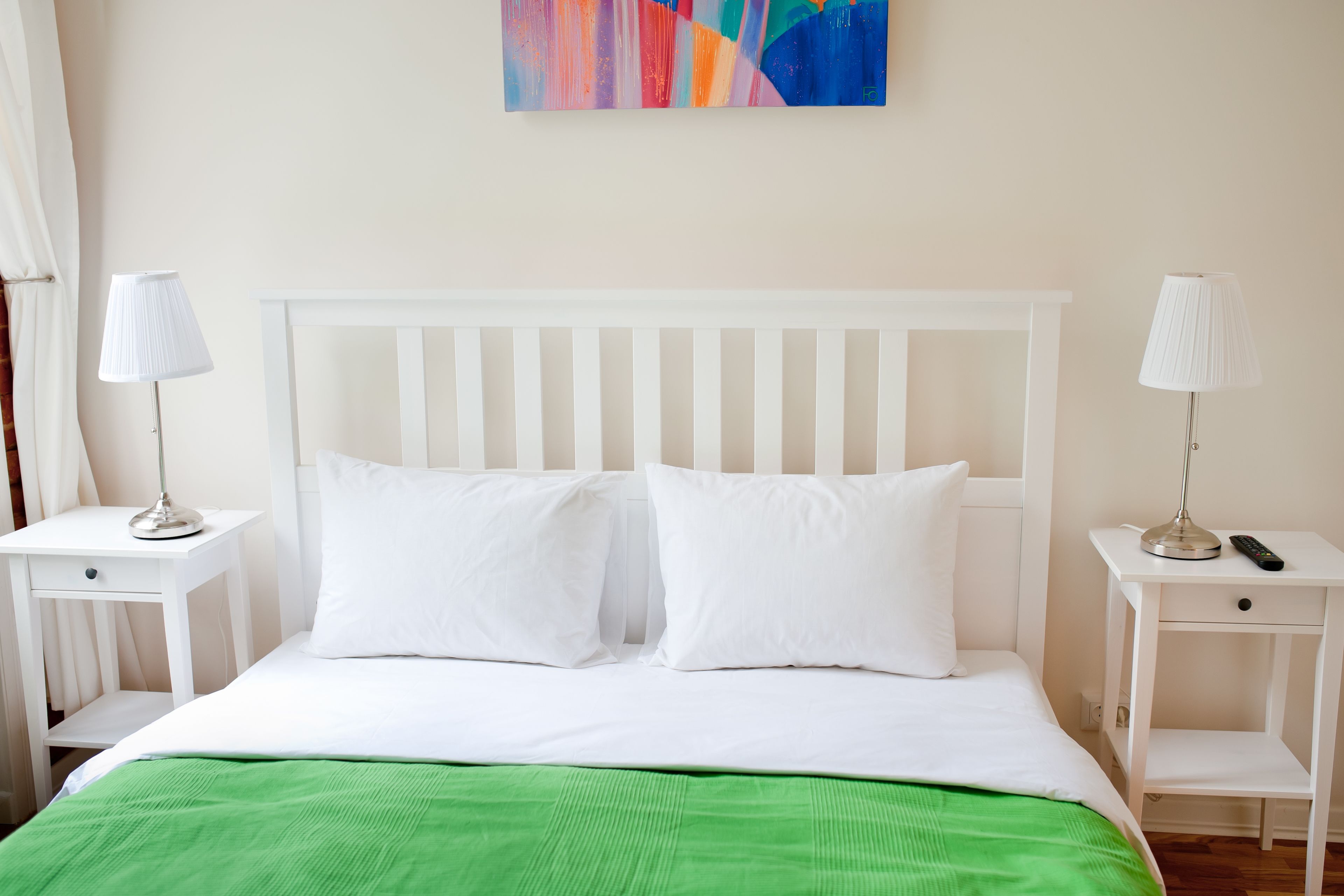 A neatly made bed with white bedding and a green throw blanket, flanked by white nightstands with lamps and a remote control.