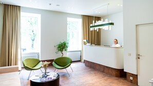 A reception area with a reception desk, two green chairs, a vase with flowers, and a large window with curtains.