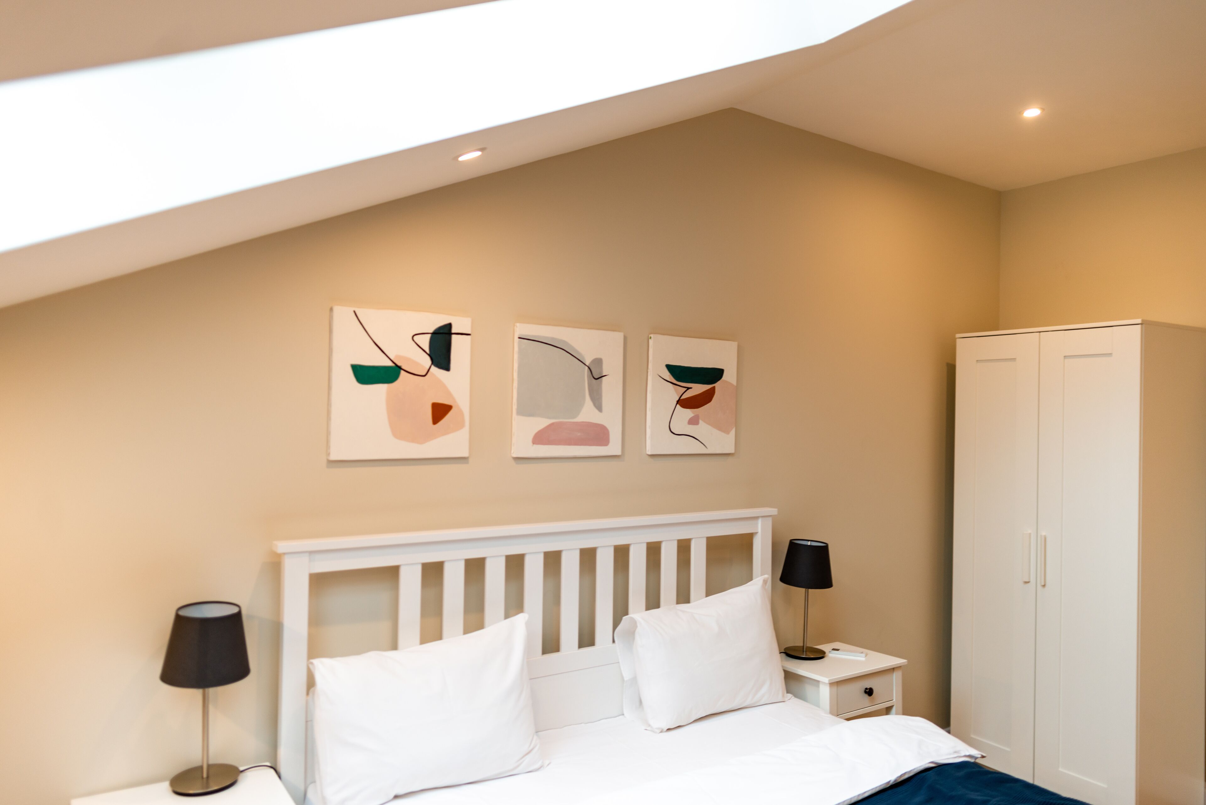 A bedroom with a white bed, bedside tables with lamps, a wardrobe, and abstract wall art.