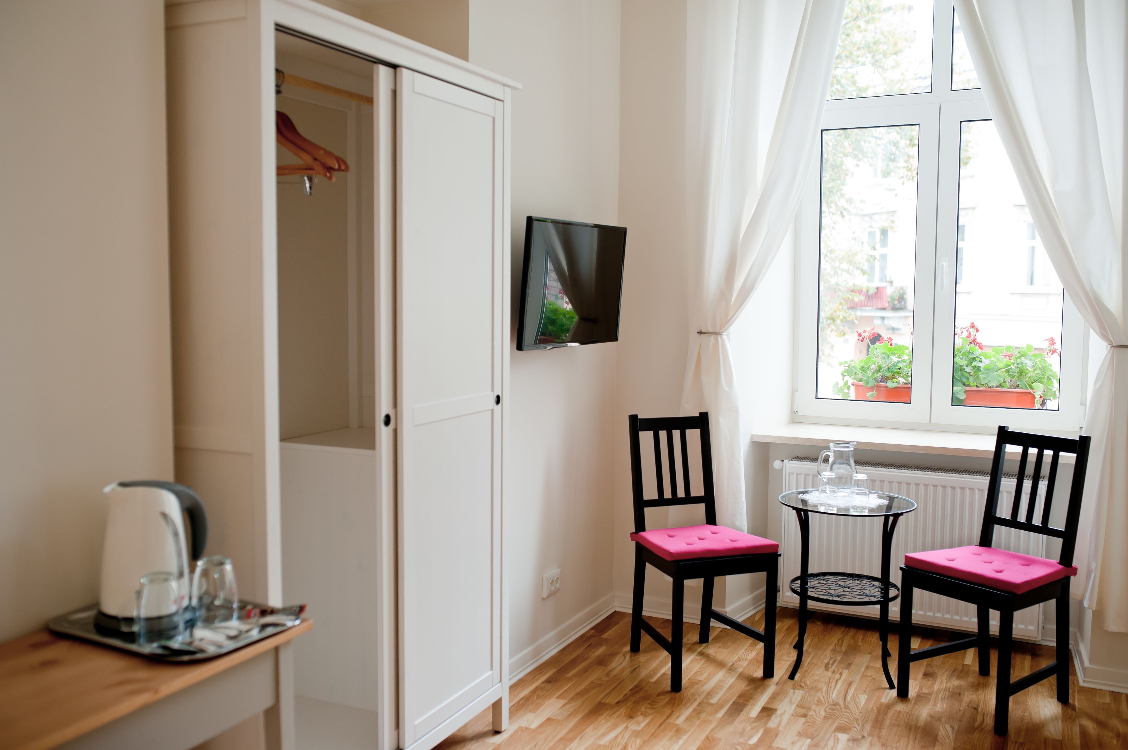 A room with a white wardrobe, a flat-screen TV mounted on the wall, two black chairs with pink cushions, a small round table, and a window with curtains.