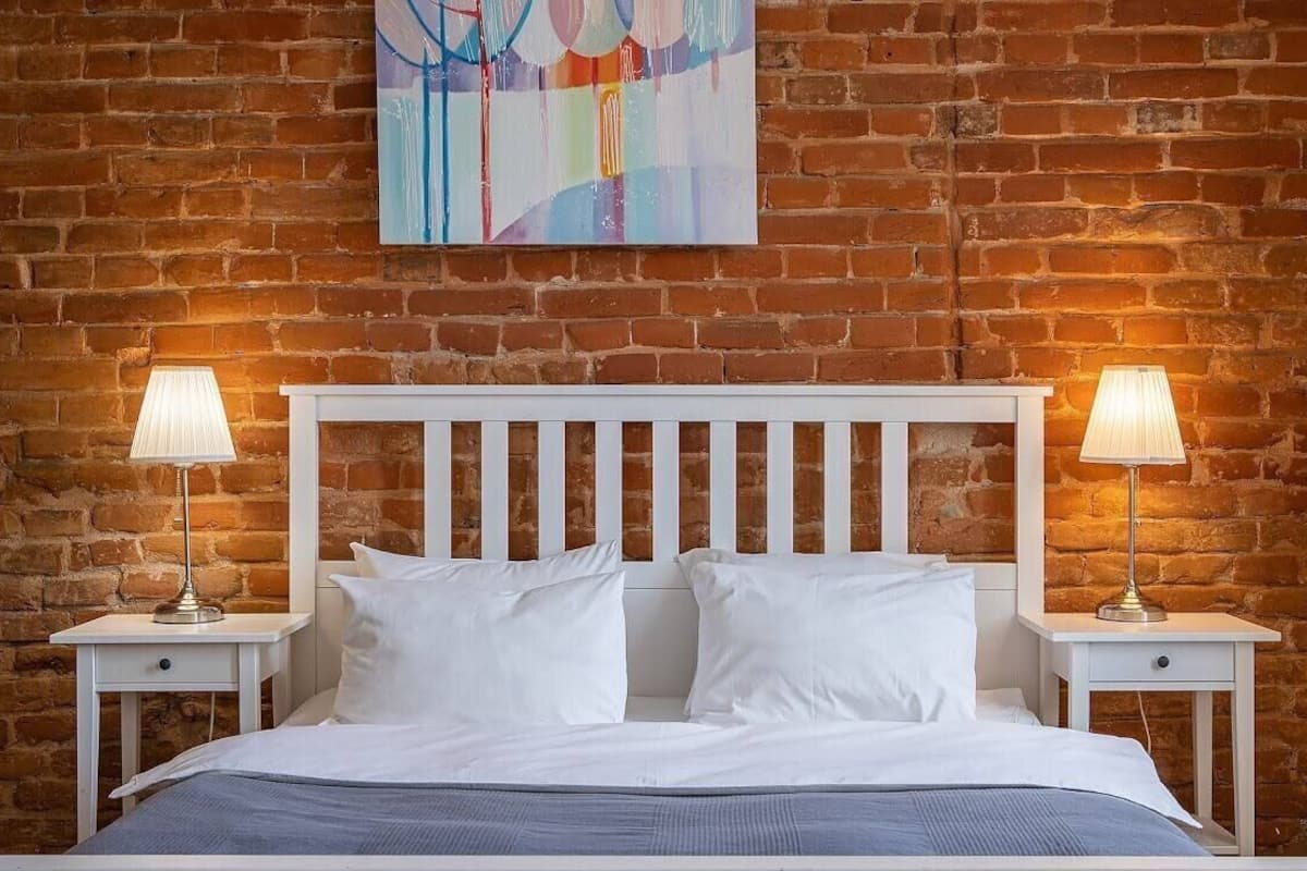 A bedroom with a brick wall, a white bed, two bedside tables with lamps, and a colorful abstract painting above the bed.