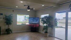A lobby with a ceiling fan, a television displaying a commercial, and a view of a parking lot through large windows. - Crossroads Inn & Suites (Victoria)