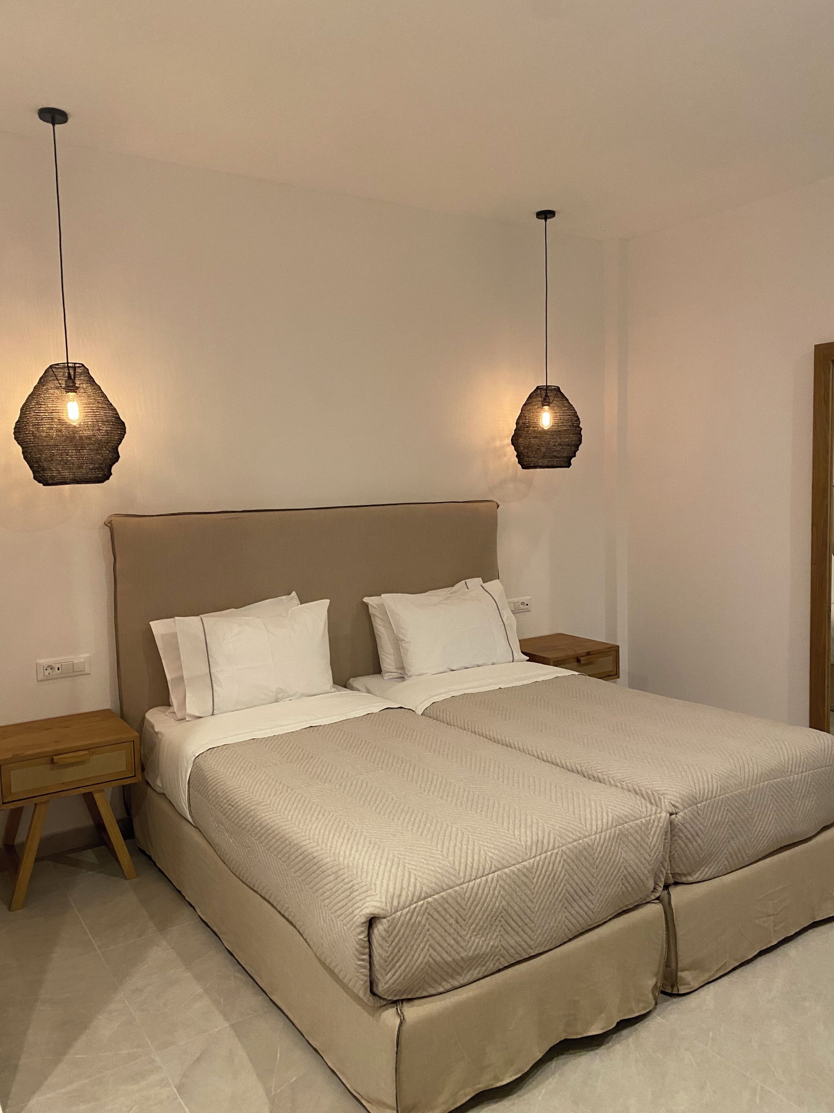 En dubbelsäng med en beige sänggavel och vita sängkläder, två sängbord med trädetaljer och två hängande taklampor med en strukturerad design.