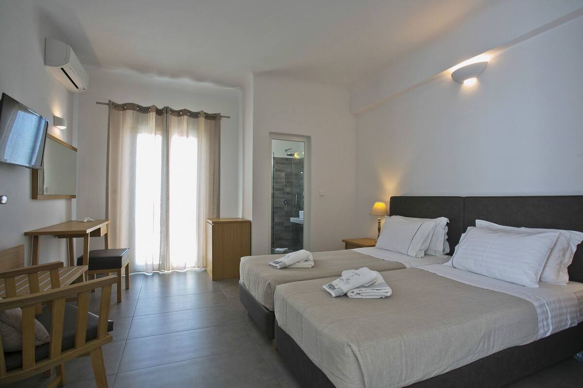 A hotel room with two beds, a desk, a chair, a TV, and a shower area.