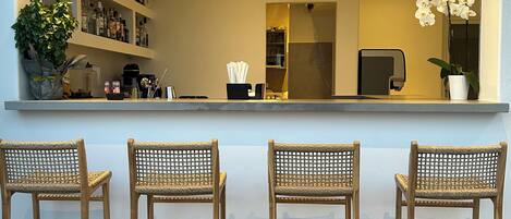 A bar area with three wicker chairs, a shelf with bottles, and a potted plant.