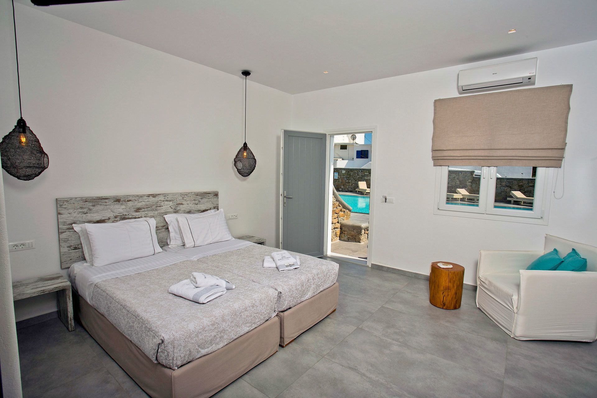 Camera da letto moderna con un letto grande, una testiera in legno, un comodino, una sedia, uno sgabello di legno e vista su una piscina attraverso una porta aperta.