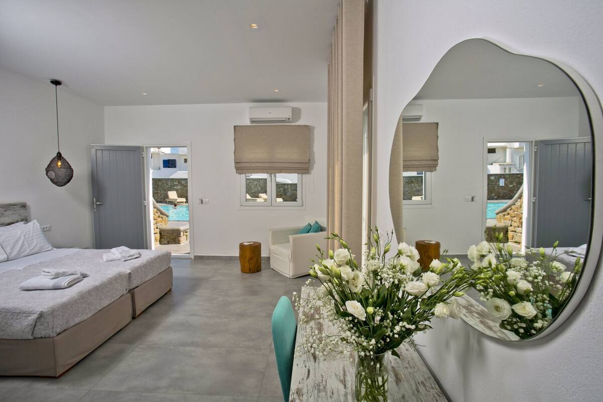 A modern bedroom with a large mirror, a bed with a white duvet, a bedside table with a vase of flowers, and a view of a pool through the door.