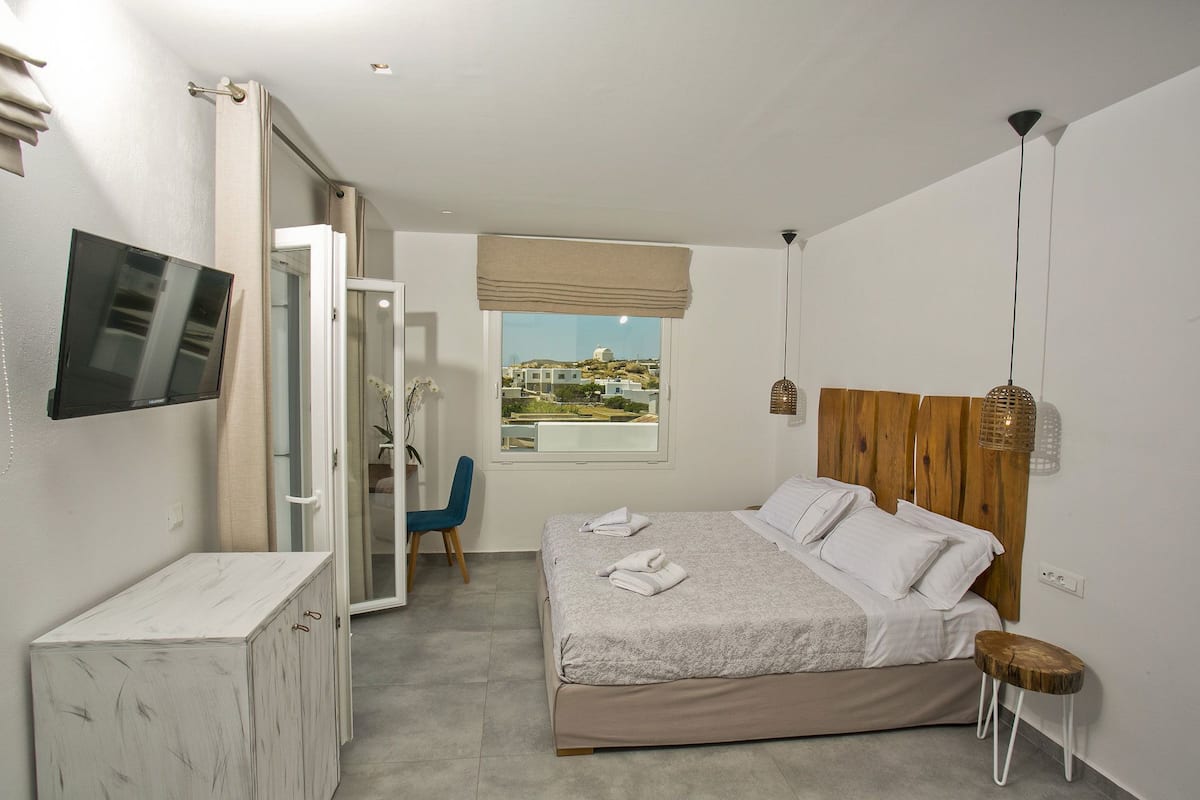 A bedroom with a bed, a TV mounted on the wall, a wooden headboard, a small wooden stool, and a view of the outside.