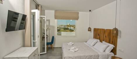 A bedroom with a bed, a TV mounted on the wall, a wooden headboard, a small wooden stool, and a view of the outside.