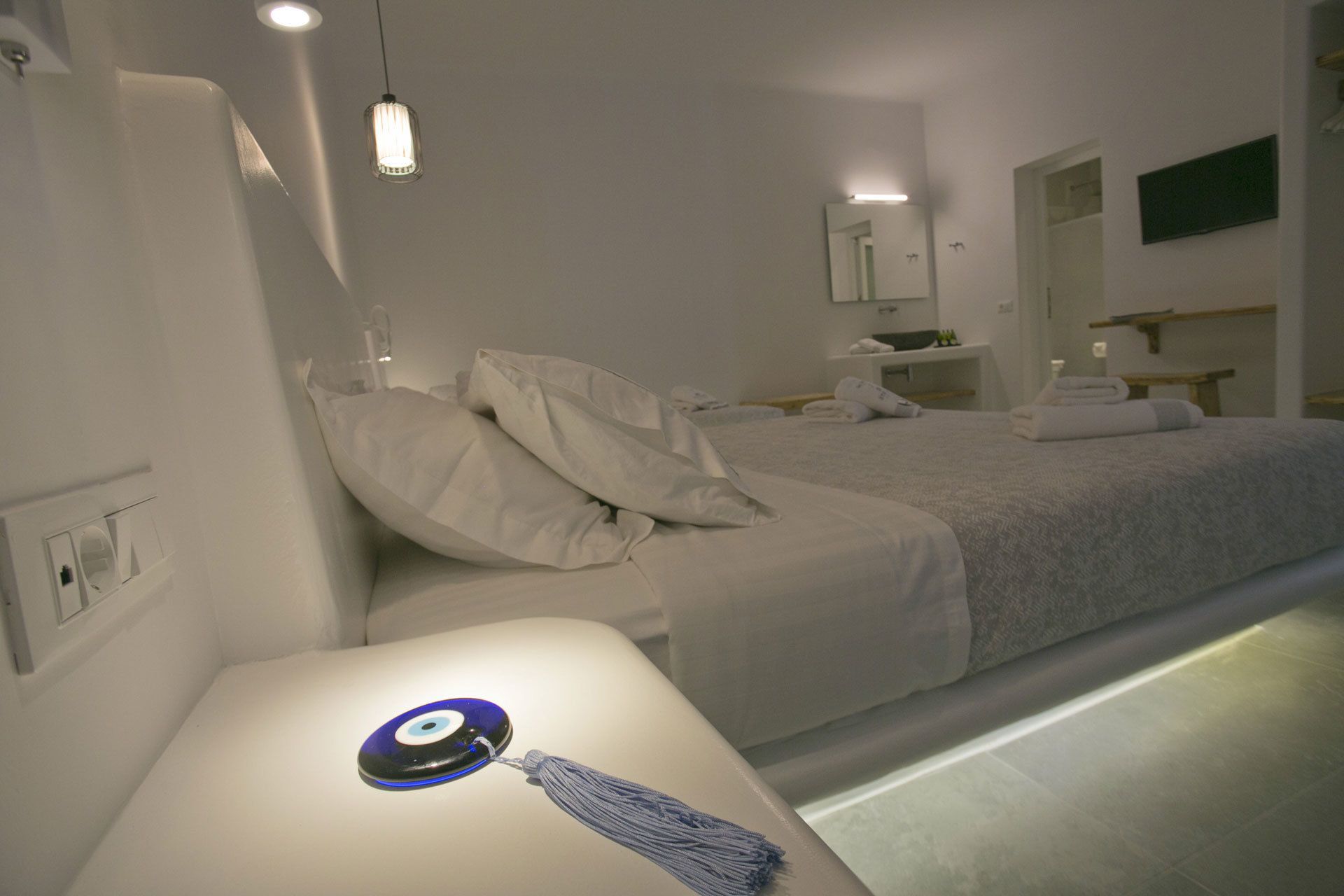 Una habitación de hotel moderna con una cama bien hecha, una mesita de noche con un objeto decorativo y un baño a la vista al fondo.