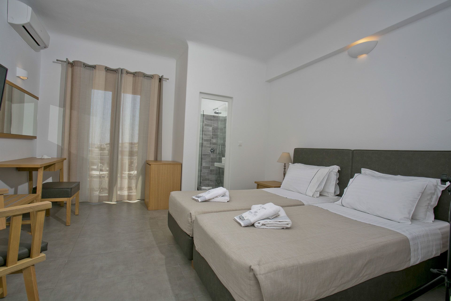 A hotel room with two beds, a desk, a chair, a wardrobe, a shower area, and a window with curtains.
