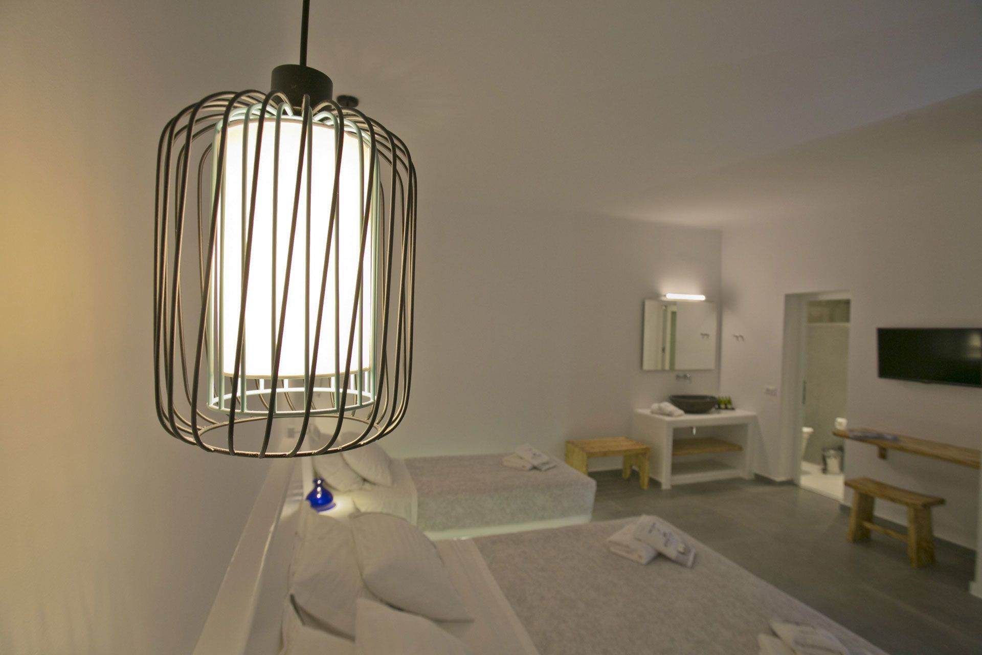 A modern hotel room with a minimalist design, featuring a white bed, a small bedside table, a built-in bench, and a large mirror above the sink.