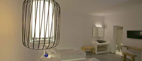 A modern hotel room with a minimalist design, featuring a white bed, a small bedside table, a built-in bench, and a large mirror above the sink.