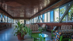Un espace extérieur abrité avec des chaises et des tables vertes, de grandes fenêtres et une plante en pot.