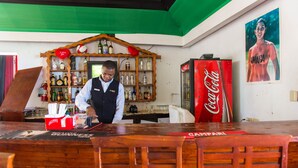 Un coin bar avec un barman, des étagères remplies de bouteilles d’alcool, un réfrigérateur à Coca-Cola et une affiche représentant un athlète jamaïcain.