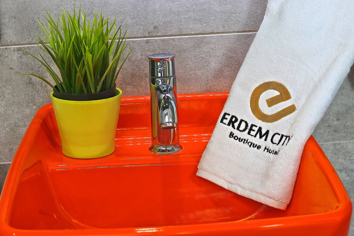 Ein Badezimmerwaschbecken mit chromfarbenem Wasserhahn, eine gelbe Pflanze in einem gelben Topf und ein weißes Handtuch mit dem Aufdruck 'ERDEM CITY Boutique Hotel'.